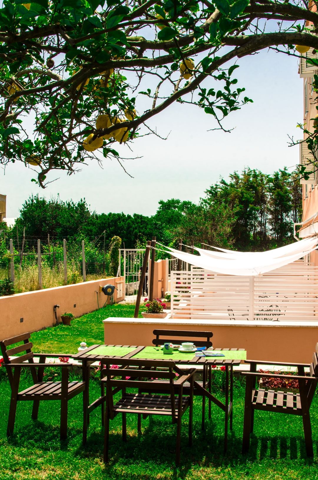 A peaceful outdoor dining area with black wooden furniture under a large tree, overlooking manicured gardens and countryside.
