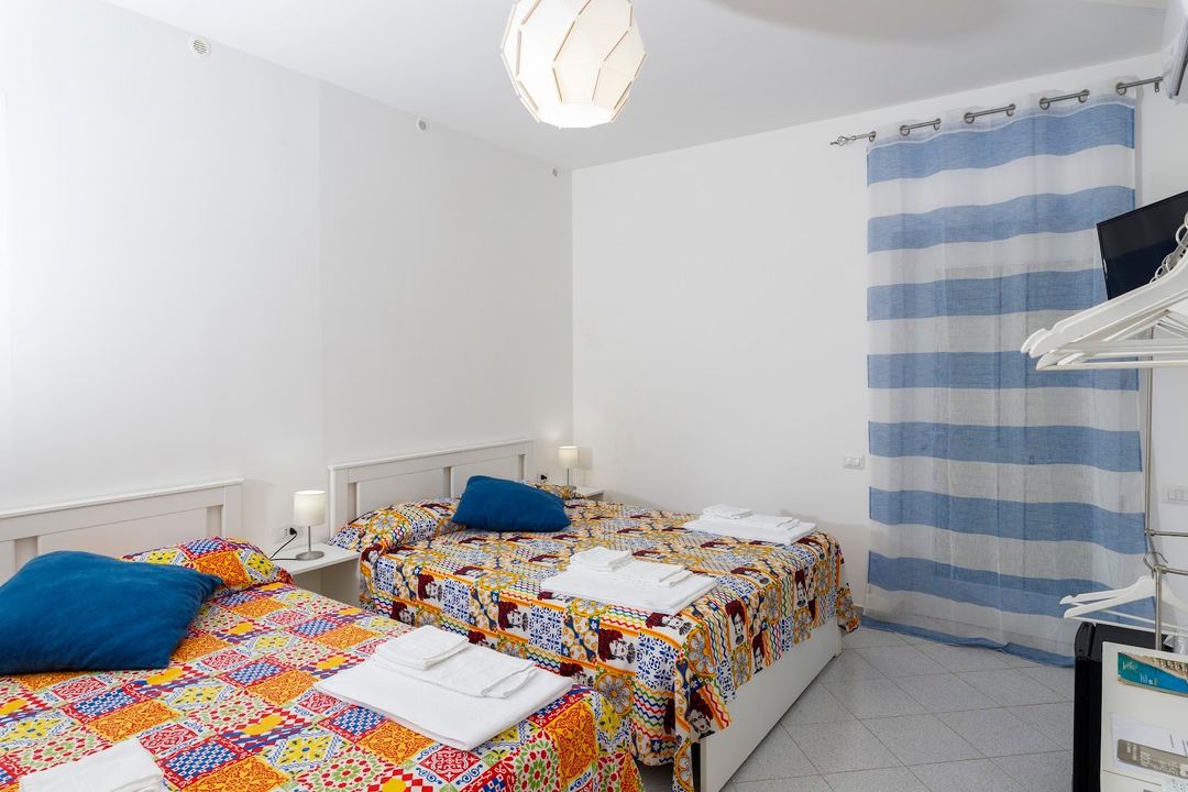 A bright hotel bedroom with two twin beds featuring colorful patchwork bedding, blue accent pillows, and a blue and white striped curtain.