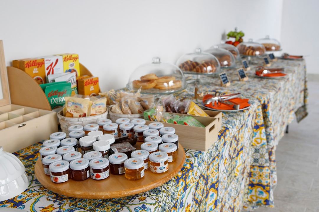 A breakfast buffet display featuring jams in glass jars, cereals, breads, and covered serving dishes arranged on a decoratively patterned table.