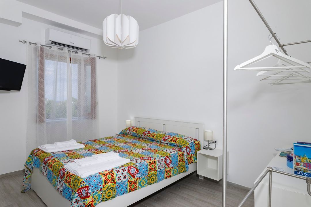 A bright, minimalist bedroom with a colorful patchwork quilt, white furniture, and modern pendant lighting.