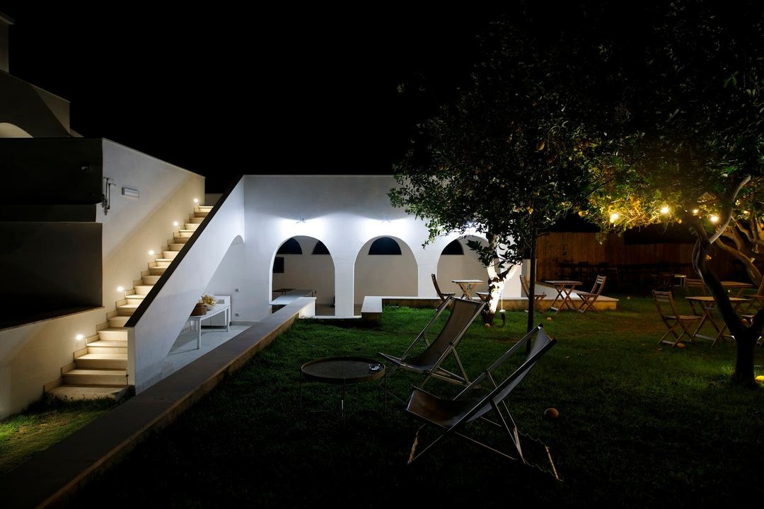 Nighttime view of a modern white building with arched openings and illuminated architectural details, overlooking a manicured lawn with lounge chairs and ambient tree lighting.