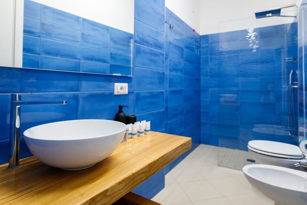 Modern bathroom with vibrant blue tile walls, a white vessel sink on a wooden vanity, and a glass-enclosed shower area.