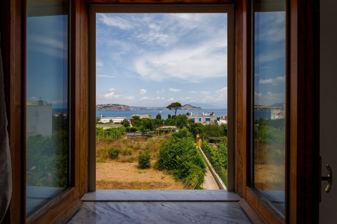Panoramic Mediterranean sea view framed through open wooden doors, overlooking a coastal property with islands and sailboats in the distance.